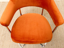 Load image into Gallery viewer, An orange velvet armchair with a curved backrest and seat, supported by slim metallic legs, photographed from a top-down angle on a textured beige woven rug.

