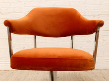 Load image into Gallery viewer, An orange velvet armchair with a curved backrest and chrome metal legs, set against a white brick wall background.
