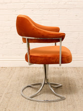 Load image into Gallery viewer, A mid-century modern swivel chair with orange velvet upholstery and a polished chrome metal base, set against a white brick wall and light beige rug.
