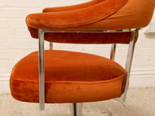 Load image into Gallery viewer, Close-up of a mid-century modern armchair with orange velvet upholstery and a polished chrome metal frame, set against a white brick wall background.
