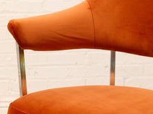 Load image into Gallery viewer, Close-up of an orange upholstered chair with a curved backrest and chrome metal legs, set against a white brick wall background.
