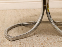 Load image into Gallery viewer, Close-up of a polished metal table base with a curved, looped leg design resting on a woven beige rug.
