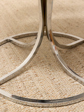 Load image into Gallery viewer, Close-up of a polished silver metal table base with curved legs resting on a woven beige rug.
