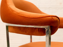 Load image into Gallery viewer, Close-up of an orange velvet upholstered chair with a curved armrest and polished chrome metal legs, set against a white brick wall background.
