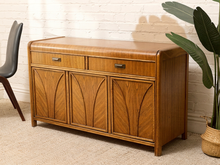 Load image into Gallery viewer, A mid-century modern wooden sideboard cabinet with two drawers and three arched cabinet doors, shown in a styled room setting with a white brick wall, beige carpet, a chair, and a potted plant.
