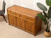 Load image into Gallery viewer, A mid-century modern wooden storage bench with a warm brown wood finish, featuring three drawers on top and three arched cabinet doors below, styled in a room corner with a potted plant and a chair.
