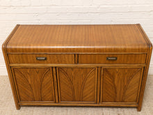 Load image into Gallery viewer, A mid-century modern wooden sideboard cabinet with a warm medium-brown finish, featuring two drawers on top and three arched cabinet doors below, set against a white brick wall and light carpet.
