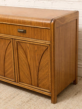 Load image into Gallery viewer, A mid-century modern wooden sideboard or credenza with a warm medium-brown finish, featuring a single drawer with a dark metal pull and two arched cabinet doors, shown in a home setting on a textured rug against a white brick wall.
