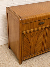 Load image into Gallery viewer, A close-up of a mid-century modern wooden sideboard or credenza with a warm medium-brown finish, featuring a single drawer with a dark metal pull and decorative carved panel doors, set against a white brick wall and beige carpet.
