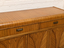 Load image into Gallery viewer, A mid-century modern wooden sideboard or credenza with a warm medium-brown wood grain finish, featuring two drawers on top with dark metal pulls and arched wood cabinet doors below, set against a white brick wall.
