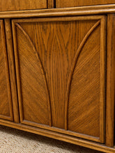 Load image into Gallery viewer, Close-up of a warm medium-brown wooden cabinet or sideboard with carved arched panel details and visible wood grain, set against a neutral beige textured surface.
