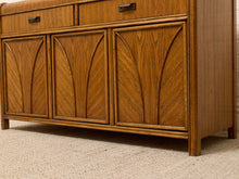 Load image into Gallery viewer, A close-up of a mid-century modern wooden sideboard or credenza with three cabinet doors featuring leaf-like carved panels and two drawers at the top, shown on a beige carpeted floor.

