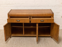 Load image into Gallery viewer, A mid-century modern wooden sideboard cabinet with a warm brown wood grain finish, featuring two open drawers on the top, open side cabinets, and open shelving below, set against a white brick wall and beige carpet.
