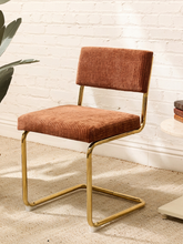 Load image into Gallery viewer, A modern accent chair with rust-brown corduroy upholstery and a curved gold metal frame, set against a white brick wall background.
