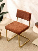 Load image into Gallery viewer, A modern accent chair with a rust-brown corduroy upholstered seat and backrest, supported by a curved gold metal frame, set on a textured beige rug against a white brick wall.
