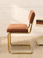 Load image into Gallery viewer, A modern cantilever dining chair with a rust-brown textured fabric seat and backrest, supported by a polished gold metal frame, set against a white brick wall and light beige rug.
