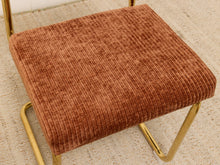 Load image into Gallery viewer, A close-up of a modern chair featuring a textured rust-brown corduroy upholstered seat and gold-finished metal legs, set on a light beige woven rug.
