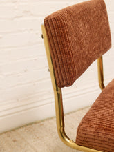 Load image into Gallery viewer, Close-up of a mid-century modern chair with a rust-brown corduroy upholstered seat and backrest, supported by a polished gold metal frame, set against a white brick wall.

