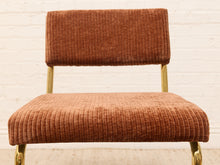 Load image into Gallery viewer, A mid-century modern accent chair with a rust-brown ribbed corduroy upholstery and gold-finished metal legs, set against a white brick wall background.
