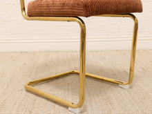 Load image into Gallery viewer, A close-up of a mid-century modern chair featuring a polished gold cantilever frame and a rust-brown corduroy upholstered seat cushion, set on a beige textured rug against a white brick wall.
