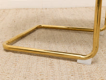 Load image into Gallery viewer, A close-up of a polished gold metal side table leg frame resting on a textured beige woven rug.
