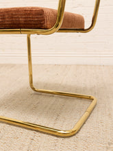 Load image into Gallery viewer, Close-up of a cantilever chair with a polished gold metal frame and a brown corduroy upholstered seat cushion, resting on a textured beige rug against a white brick wall.
