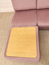 Load image into Gallery viewer, A mauve-pink modular sofa with a light wood tray table attached to the front of one seat cushion, set on a textured beige rug.
