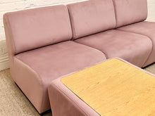 Load image into Gallery viewer, A dusty pink velvet modular sofa with a matching ottoman featuring a light wood-grain top, set against a white brick wall and beige carpet.
