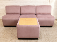 Load image into Gallery viewer, A three-seat modular sofa in dusty pink velvet with a matching ottoman featuring a light wood top, set against a white brick wall and beige rug.
