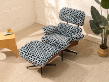 Load image into Gallery viewer, A mid-century modern lounge chair with an attached ottoman, upholstered in a navy, gray, and white geometric patterned fabric, set in a sunlit room with a side table and potted plant.
