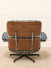 Load image into Gallery viewer, A mid-century modern lounge chair with a curved wooden shell, black metal swivel base, and black-and-white geometric patterned fabric upholstery, set against a white brick wall and beige rug.
