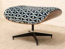 Load image into Gallery viewer, A mid-century modern lounge ottoman with a curved wooden shell, black metal star base, and textured blue and black patterned fabric upholstery, set on a beige carpet against a white brick wall.
