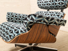 Load image into Gallery viewer, A close-up of a mid-century modern lounge chair with patterned blue, black, and white fabric upholstery and a curved wooden shell, shown in a styled interior setting.
