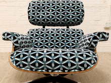 Load image into Gallery viewer, An upholstered lounge chair with a black, blue, and white geometric patterned fabric, featuring a wooden armrest frame and a black swivel base, set against a white brick wall background.
