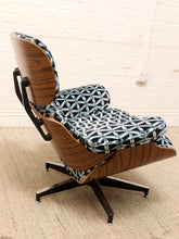 Load image into Gallery viewer, A mid-century modern lounge chair with a curved dark wood shell, black metal swivel base, and patterned blue, black, and white seat and back cushions, set against a white brick wall and beige carpet.
