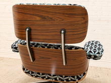 Load image into Gallery viewer, A mid-century modern lounge chair with a curved wooden shell, dark wood grain backrest, and patterned black, white, and blue fabric upholstery, shown against a white brick wall.
