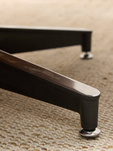 Load image into Gallery viewer, Close-up of a dark wood or metal furniture leg with a silver round caster wheel, resting on a textured beige woven rug.

