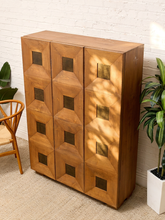 Load image into Gallery viewer, A mid-century modern wooden cabinet with a geometric, faceted triangular pattern on the front and dark square metal pulls, set against a white brick wall with plants and a chair.
