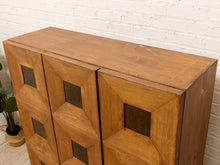Load image into Gallery viewer, A mid-century modern wooden cabinet with geometric inset panels and dark square glass or metal inserts, set against a white brick wall.
