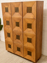 Load image into Gallery viewer, A mid-century modern wooden cabinet with a warm honey finish, featuring a geometric pattern of raised diamond-shaped panels and square recessed details on the doors, set against a white brick wall and beige carpet.
