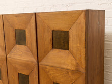 Load image into Gallery viewer, A close-up of a mid-century modern wooden cabinet featuring geometric, stepped panel designs and square recessed details, set against a white brick wall.
