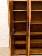 Load image into Gallery viewer, A tall wooden bookcase with open doors and multiple shelves, shown against a white brick wall and light carpet.
