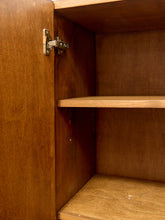 Load image into Gallery viewer, Close-up of the inside of a wooden cabinet with one door open, showing a metal hinge, a middle shelf, and a bottom shelf against warm brown wood grain.
