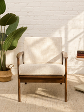 Load image into Gallery viewer, A mid-century modern accent chair with a light beige textured linen cushion and a medium brown wooden frame, set against a white brick wall with a potted green plant on the left.
