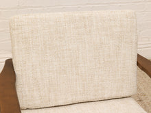 Load image into Gallery viewer, A close-up of a beige textured fabric chair cushion resting on a wooden armchair against a white brick wall background.
