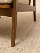 Load image into Gallery viewer, Close-up of the lower portion of a wooden chair with a beige upholstered seat, standing on a textured cream woven rug.
