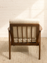 Load image into Gallery viewer, A mid-century modern wooden armchair with a warm brown frame and textured beige fabric upholstery, shown from the back against a white brick wall and light-colored rug.
