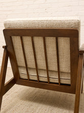 Load image into Gallery viewer, Close-up of the back of a mid-century modern armchair with a dark brown wooden frame, vertical slats, and textured beige fabric upholstery, set against a white brick wall.
