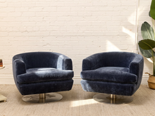 Load image into Gallery viewer, Two navy blue velvet swivel armchairs with round metallic bases, positioned side-by-side in a bright room with a white brick wall and a potted plant.
