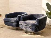 Load image into Gallery viewer, Two modern barrel-style swivel armchairs upholstered in dark blue velvet, set on round brushed metal bases against a white brick wall.

